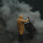High-End - A man in a yellow rain coat standing on a cliff with smoke coming out of the water