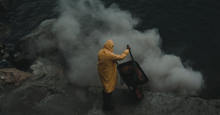 High-End - A man in a yellow rain coat standing on a cliff with smoke coming out of the water