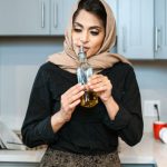 Odor - Positive stylish ethnic female in headscarf smelling aromatic olive oil in bottle while cooking dinner in contemporary light kitchen