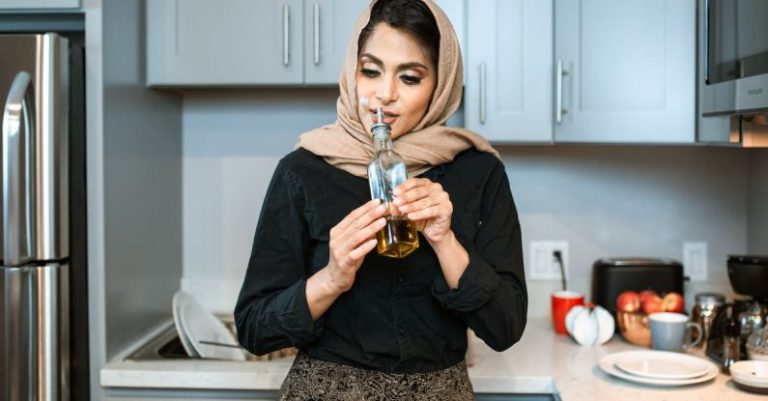 Odor - Positive stylish ethnic female in headscarf smelling aromatic olive oil in bottle while cooking dinner in contemporary light kitchen