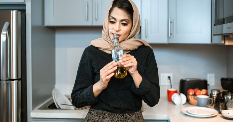Odor - Positive stylish ethnic female in headscarf smelling aromatic olive oil in bottle while cooking dinner in contemporary light kitchen